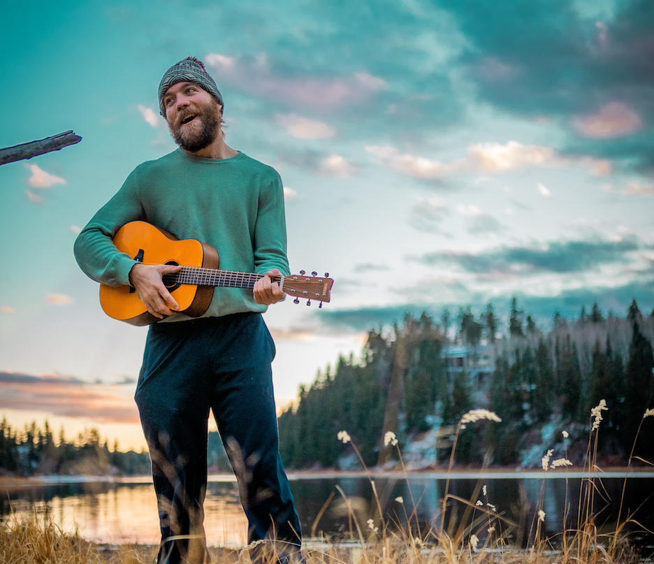 Man with a beard wearing a green beanie hat, sweater, and jeans strumming a guitar in front of a lake.