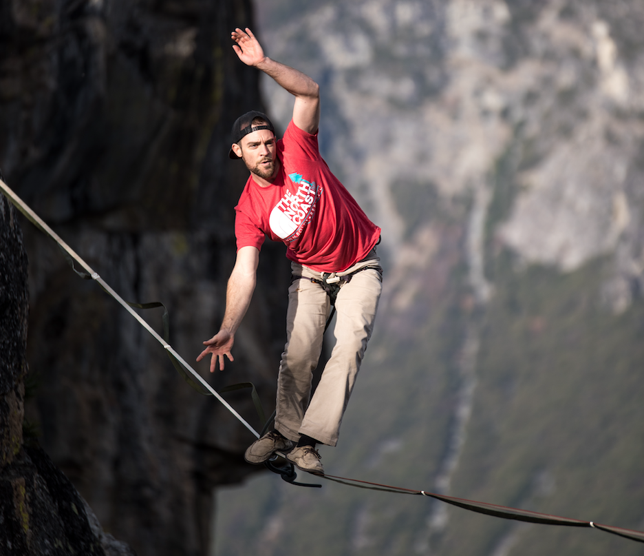 Man wearing a red shirt walking a tight rope (technically a slack line) high above a cliff